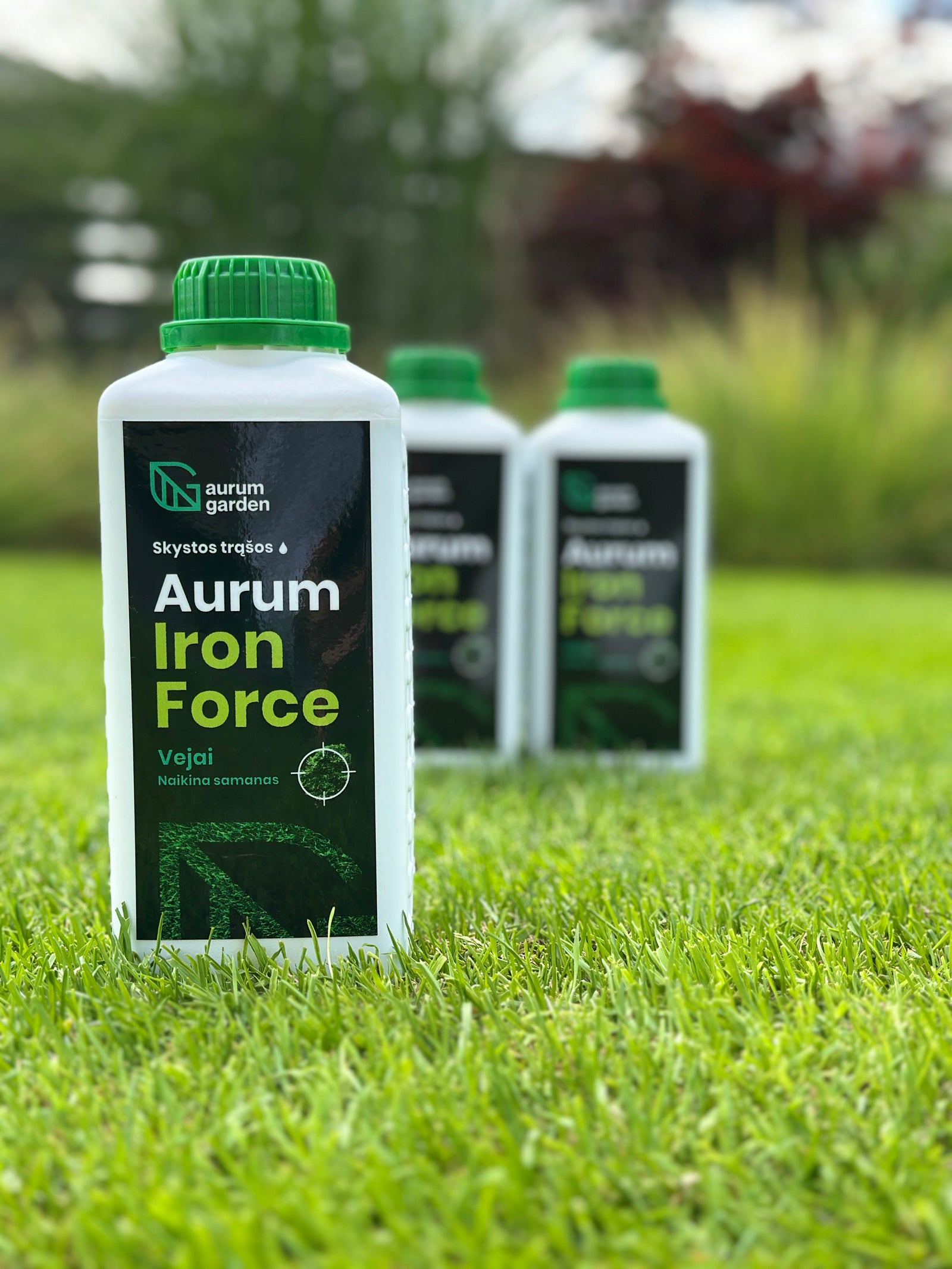 „Aurum Iron Force“ trąšų pakuotė stovi vejoje. Fone neryškiai matomos kitos produkto pakuotės ir sodo augalai.