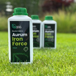 „Aurum Iron Force“ trąšų pakuotė stovi vejoje. Fone neryškiai matomos kitos produkto pakuotės ir sodo augalai.